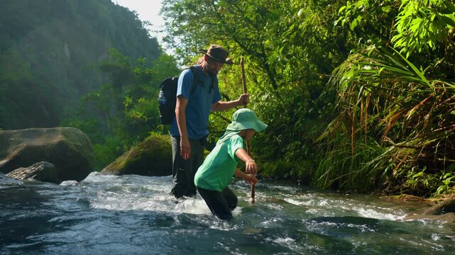 Father and son wade through flowing tropical river in lush green jungle setting.