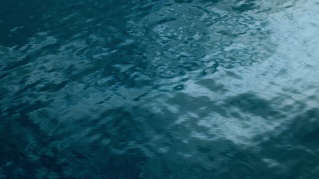 Gentle raindrops fall onto a calm blue water surface creating delicate circular ripples that spread outward. The deep teal water reflects soft light as rain continues to disturb the tranquil scene.