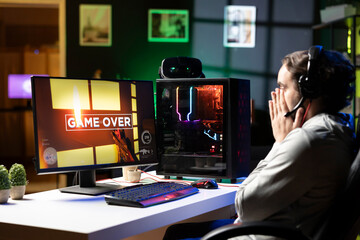 Sad gamer upset after losing online multiplayer action videogame championship, being defeated by rivals. Man putting face in palms in frustration after seeing game over screen on gaming pc display © DC Studio