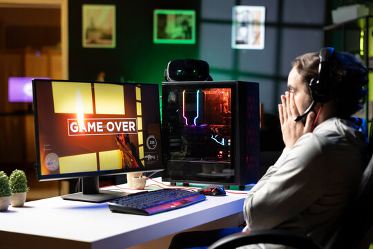 Sad gamer upset after losing online multiplayer action videogame championship, being defeated by rivals. Man putting face in palms in frustration after seeing game over screen on gaming pc display