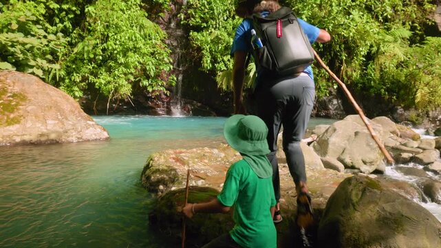 Father and young son in green hat carefully navigate mossy boulders beside crystal clear turquoise river pool with gentle waterfall cascading through dense tropical rainforest foliage in background.