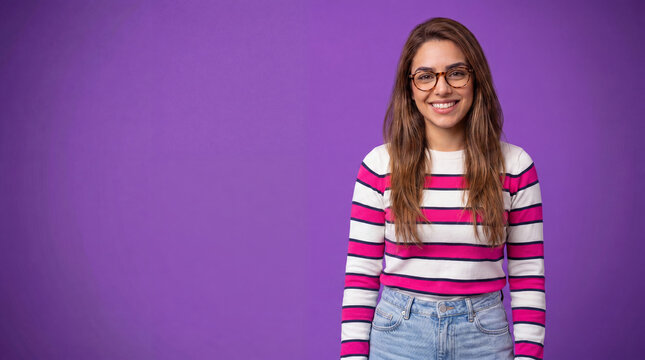 A young woman with glasses and long brown hair, wearing a striped sweater and jeans, smiles warmly at the camera against a vibrant purple background