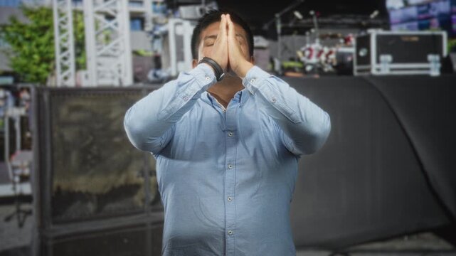 Man in light blue button shirt pressing bare hands together in prayer and looking upward on a crowded street event stage; faith hope.