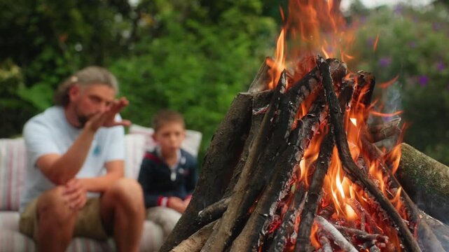 Father and son relax together by a campfire in a lush green backyard. Burning wood logs create bright orange flames and glowing embers while the man gestures during conversation with the boy.