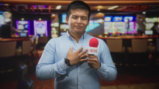 Young hispanic man reporter holding news microphone with hand on chest amid slot machines and chairs in building interior; sincerity duty.