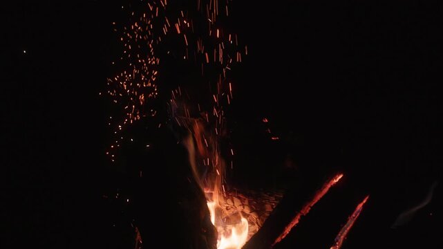 Bright orange sparks rise from a crackling campfire into the pitch black night sky. Burning wood logs glow with intense flames while embers float upward in mesmerizing patterns.