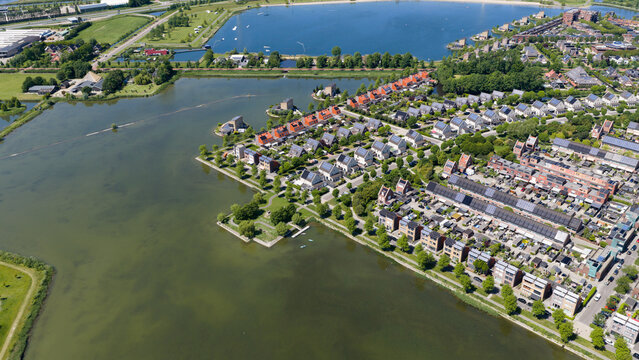 Aerial view of modern residential houses with solar panels on rooftops along a calm lake surrounded by green trees and pathways in Heerhugowaard, North Holland, Netherlands.
