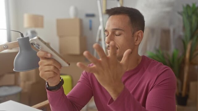 Man with smartphone holds phone and gestures with open hand while speaking, moustache visible, wearing long sleeve shirt, moving boxes and lamp around in apartment; frustration.