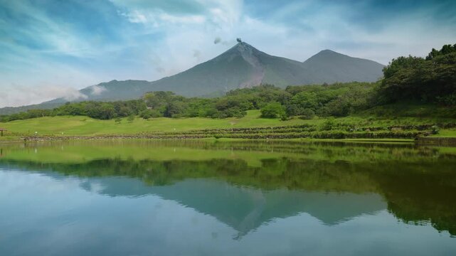 Guatemala active Fuego volcano releases smoke plumes above lush green tropical landscape. Serene lake mirrors majestic volcanic peaks and blue sky with wispy clouds creating stunning natural scenery.