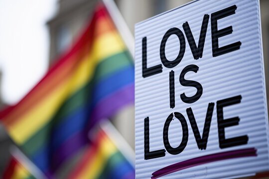 Protest sign showing text 'Love is Love' written in marker on cardboard with blurred rainbow flags in background. Generative AI, AI generated