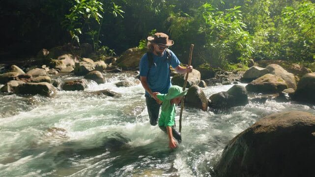 Son with father hiking through wild nature as they wade across a rushing jungle river. Dad uses walking stick to navigate rapids while guiding child past mossy boulders in lush green tropical forest.