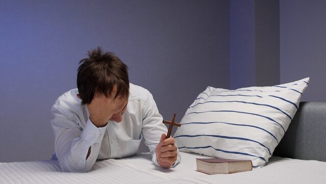 Man holding a wooden cross and praying with his head in his hand while kneeling on a bed next to a book and a striped pillow, expressing faith and seeking comfort during a difficult time
