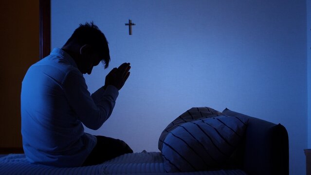Man kneeling on a bed in a dark room, silhouetted against a blue light from a window, hands clasped in prayer while a christian cross hangs on the wall