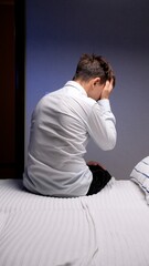 Fototapeta premium Young adult man sitting on the edge of a bed, covering his face with hands, feeling distressed and overwhelmed by sadness, depression, or a headache late at night