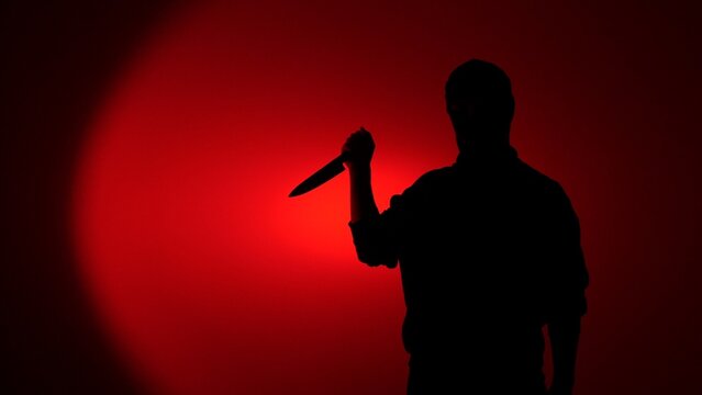 Man's silhouette appearing against a red lit background, holding a knife, creating a chilling atmosphere of fear, danger, crime, and potential violence