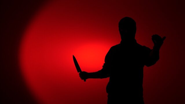 Silhouette of a threatening man holding a large kitchen knife against a red background, evoking crime, danger, suspense and a menacing, anonymous attacker in dramatic lighting