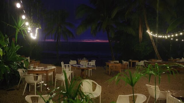 Heavy rain pours down on empty beachfront restaurant with outdoor dining tables and chairs. Palm trees sway as string lights glow against purple twilight sky over ocean horizon in tropical setting.