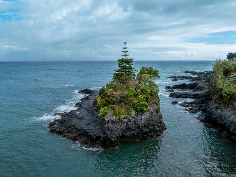 Aerial view of a rocky islet with a lone pine tree and green shrubs surrounded by the dark blue Atlantic Ocean under a cloudy sky in Caloura, Acores, Portugal.