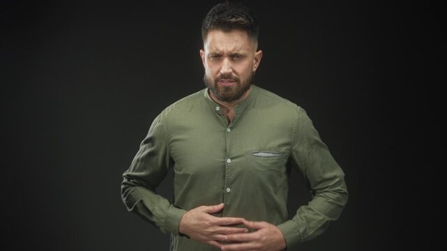 Man, bearded and young, clutching stomach with both hands, wincing and leaning forward, wearing green button shirt in studio; pain vulnerability.