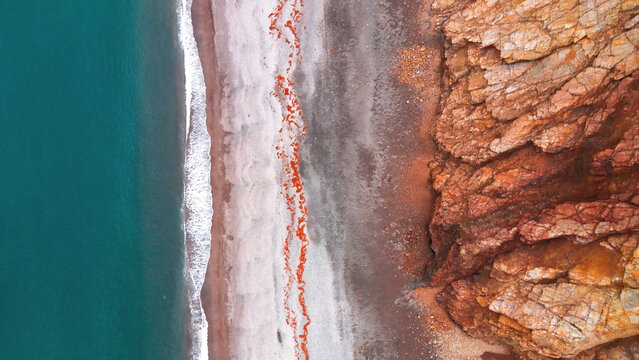Aerial view of the Brunavik coastline featuring turquoise ocean waves crashing against a dark pebble beach and steep orange cliffs in Bakkagerdi, Mulathing, Iceland.