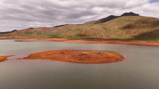Aerial view of green lake with orange nickel shores, remote Blue River Provincial Park panorama with mountains and small vegetated islands, calm water under scattered clouds. Noumea, New Caledonia.