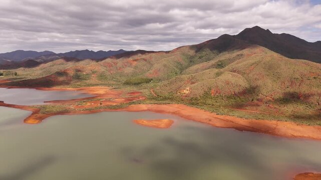 Aerial view of green lake with orange nickel shores, remote Blue River Provincial Park panorama with mountains and small vegetated islands, calm water under scattered clouds. Noumea, New Caledonia.
