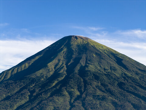 Aerial view of Mount Sindoro with its majestic conical peak and lush green slopes under a clear blue sky in Kecamatan Kertek, Jawa Tengah, Indonesia.
