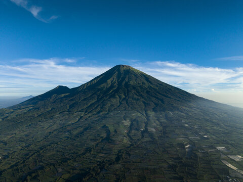 Aerial view of Mount Sindoro with its conical peak and agricultural fields on the slopes under a clear blue sky in Kecamatan Kertek, Jawa Tengah, Indonesia.