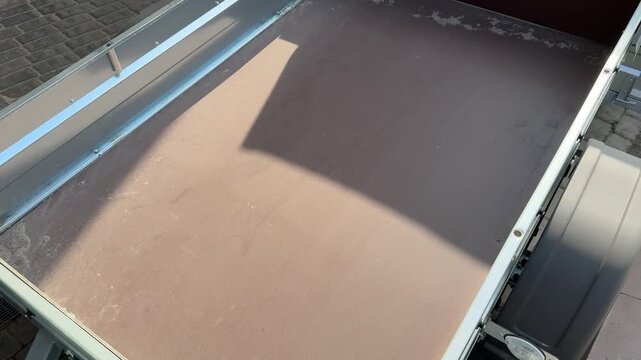 Top view empty trailer bed showing green metal frame and worn plywood floor with edge chips and small damage, sunlight casting