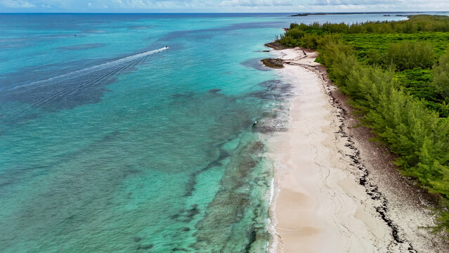 Aerial view of Preacher's cave beach with turquoise waters, white sand, and a boat leaving a wake along the lush coastline in The Bluff Settlement, North Eleuthera, The Bahamas.