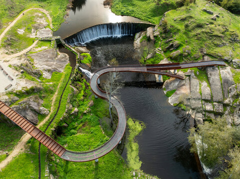 Aerial view of Acude da Ribeira weir featuring a modern sinuous metal walkway over the cascading water and green rocky riverbanks in Ervedal, Coimbra, Portugal.