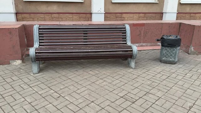 Empty park bench next to trash bin with black bag on gray paving stones during day, decorative cast iron bench ends, red low wall
