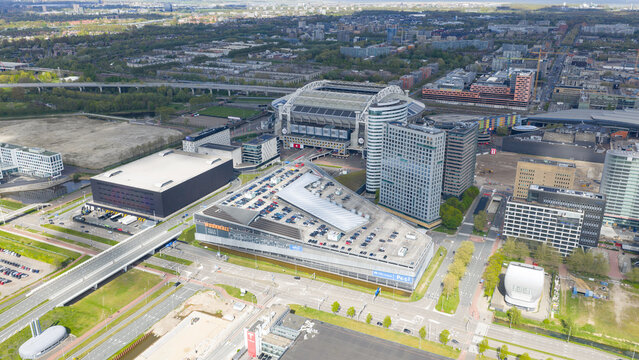 Amsterdam, Netherlands - 19 April 2026: Aerial view of the Johan Cruijff Boulevard area featuring the Johan Cruijff Arena stadium, modern office buildings, and a rooftop parking lot.