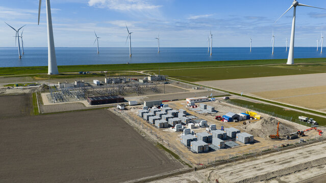 Espel, Netherlands - 04 April 2026: Aerial view of the Westermeerdijk wind farm and an electrical substation with battery storage containers near the coast under a blue sky.