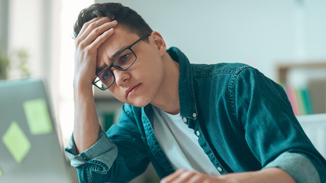 Worried Young Man With Glasses Concentrating While Working At Laptop In Modern Office Setting, Feeling Stressed About Tasks And Deadlines, Seeking Solution. Business Concept