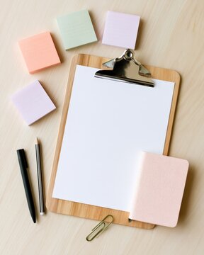 Clipboard with blank paper on wooden desk. Colorful sticky notes surround the clipboard. Two pencils and binder clip nearby. Notebook with textured cover lies beside. Scene suggests planning