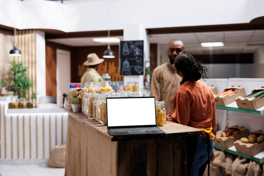 Placed on a wooden counter is a digital laptop showing an isolated white screen display. A wireless computer at a contemporary grocery shop has a blank chromakey mockup template.