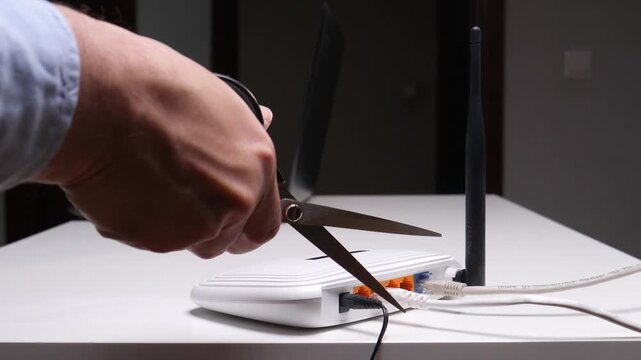 Person's hand holding open scissors, preparing to cut an ethernet cable plugged into a white wireless broadband router, symbolizing disconnection from internet access
