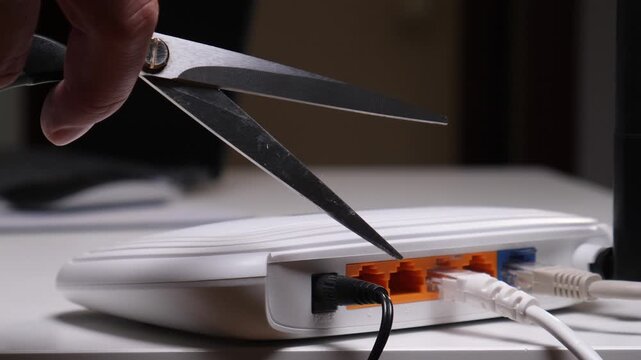 Person's hand holding open scissors, positioned to cut a cable plugged into a white internet router, symbolizing digital disconnection, censorship, or a break from online activity