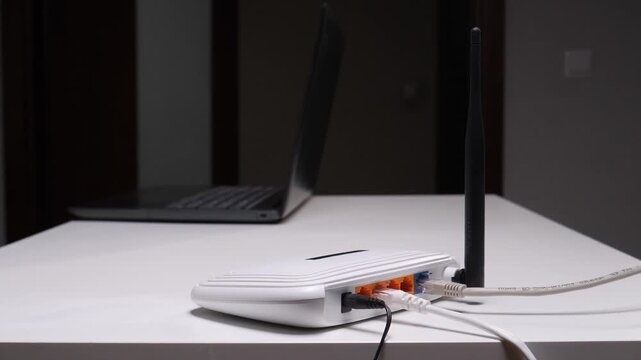 Man's hands plugging an ethernet cable into a white wireless broadband wifi router on a white desk with a laptop in the blurry background, setting up internet connection for home or office