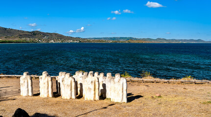 Obraz premium The Cap 110 Memorial at Anse Caffard in Martinique features white stone statues overlooking the blue Caribbean Sea and coastal hills.