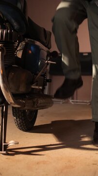 Vertical video person in boots and work pants kickstarting a vintage motorcycle in a dim garage, focusing on the engine and kick lever as they prepare the classic bike for startup