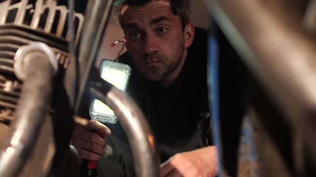 Man shining a flashlight onto a motorcycle engine, carefully inspecting components during maintenance or repair work, demonstrating expertise and focus in a professional garage setting