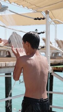 Shirtless boy rinsing off under outdoor beach shower near sea after swimming. Vertical reel of sunny family resort vacation, summer travel, hygiene and water lifestyle concept. Beach shower on pier