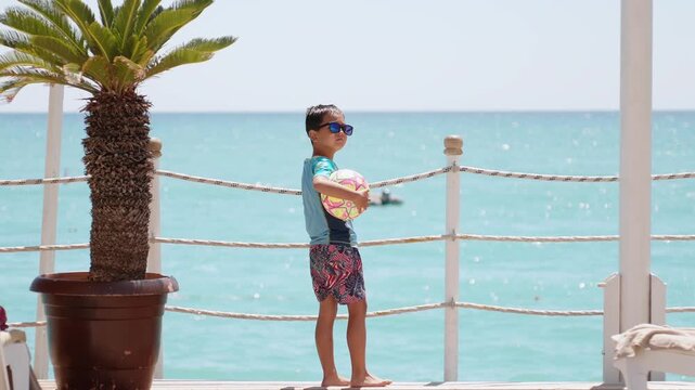 Young boy wearing sunglasses and holding colorful beach ball near turquoise sea. Sunny tropical resort vacation, family travel and summer leisure lifestyle