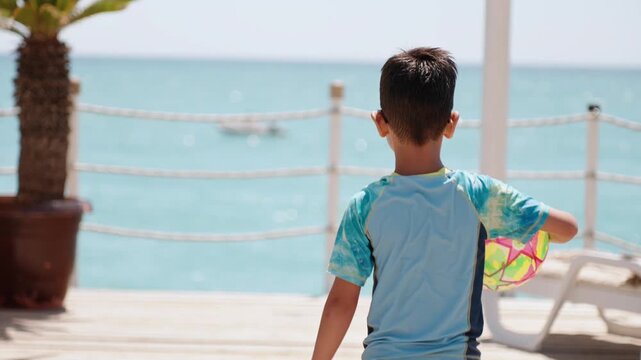 Young child standing barefoot with colorful beach ball on wooden seaside deck, looking at sea during sunny family resort holiday and summer travel. Kid holding ball heads to the edge of pier
