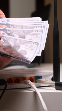 Vertical video hand holding hundred dollar bills above a wireless internet router with connected network cables, visualizing the recurring costs for broadband service and technology expenses
