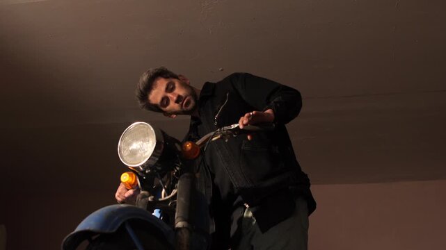 Man in black jacket inspects handlebars and headlight of vintage blue motorcycle in a dim garage, focused on repair and maintenance with professional, skilled hands