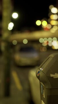 A parking meter is visible on a city street at night surrounded by cars and soft lights glowing in the background. The scene shows a typical urban atmosphere with no people.