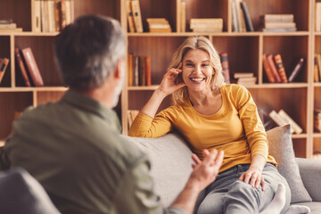 Fototapeta premium Smiling Caucasian Woman Engaging In Friendly Conversation With A Man While Sitting On Sofa In Cozy Living Room, Enjoying Each Other's Company. Socializing Concept